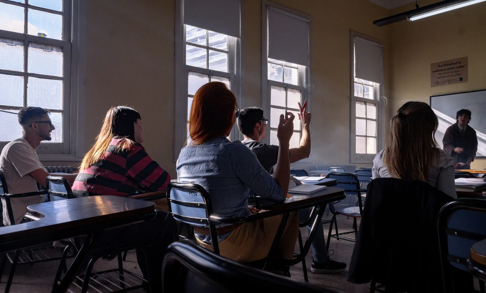 Estudiantes de espaldas atienden una clase en una sala con ventanales; uno levanta la mano.
