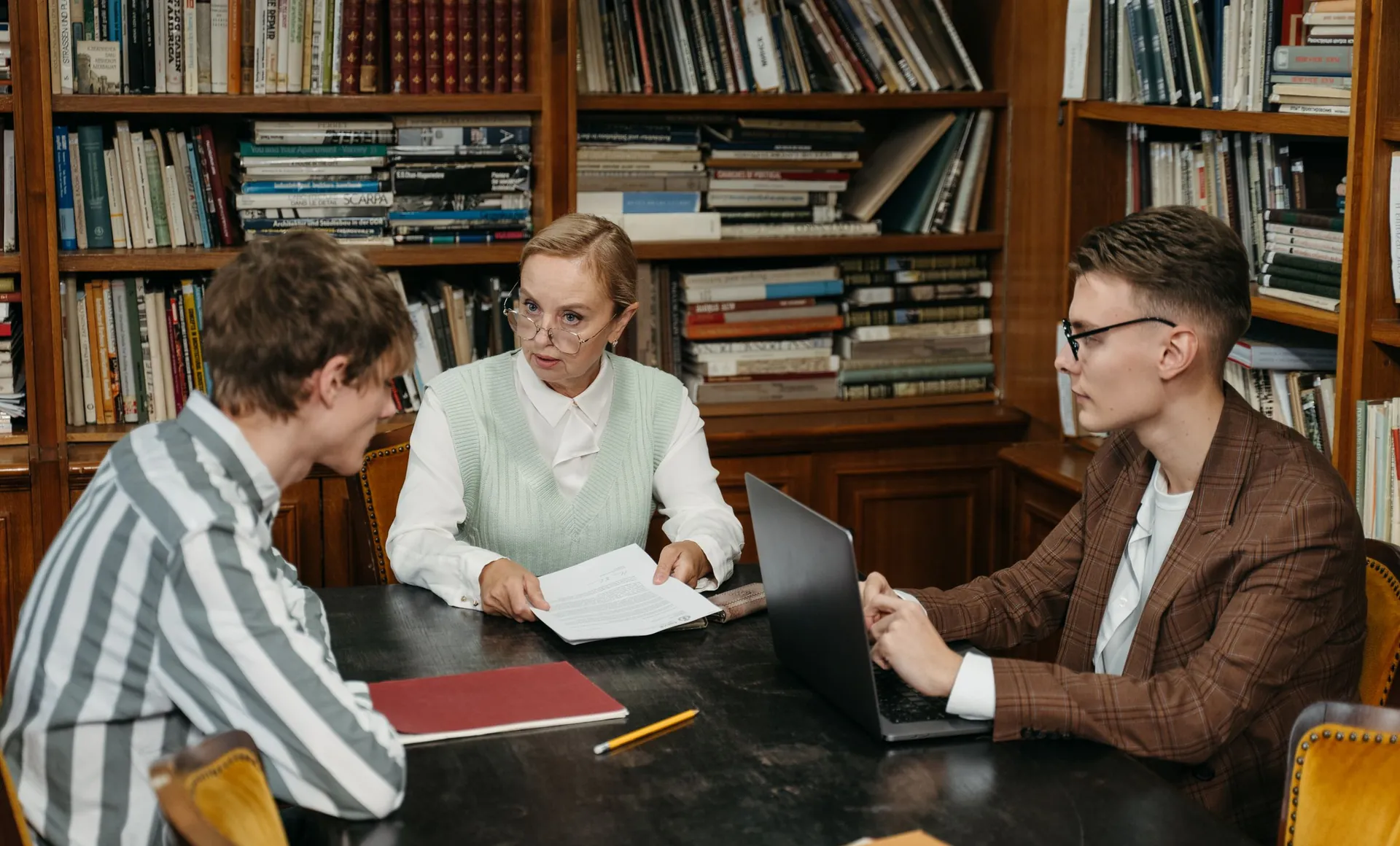 Tres personas revisan documentos sobre una mesa en una oficina con estantes de libros.